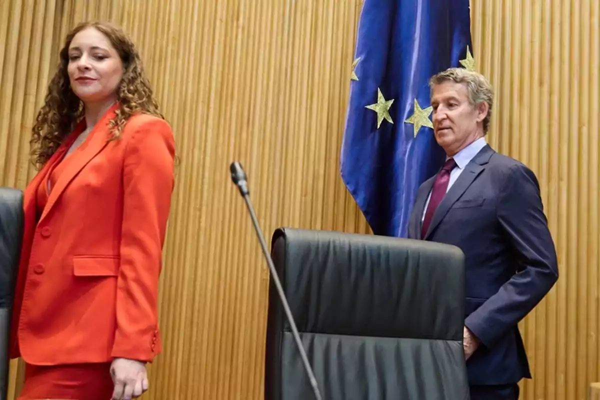 Una mujer con traje rojo y un hombre con traje oscuro están de pie en una sala con paredes de madera y una bandera azul con estrellas amarillas al fondo Una mujer con traje rojo y un hombre con traje oscuro están de pie en una sala con paredes de madera y una bandera azul con estrellas amarillas al fondo