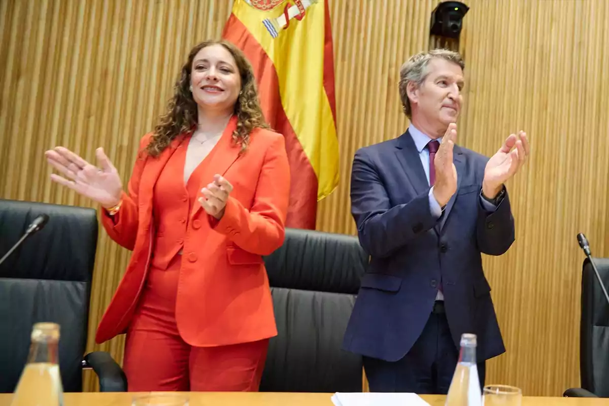 Dos personas aplauden de pie en una sala de conferencias con una bandera de España al fondo