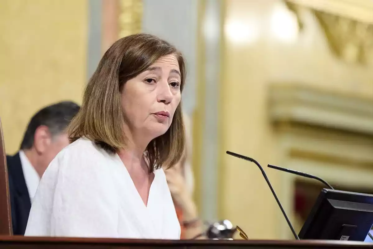 Mujer de cabello castaño y ropa blanca hablando frente a un micrófono en un entorno institucional Mujer de cabello castaño y ropa blanca hablando frente a un micrófono en un entorno institucional