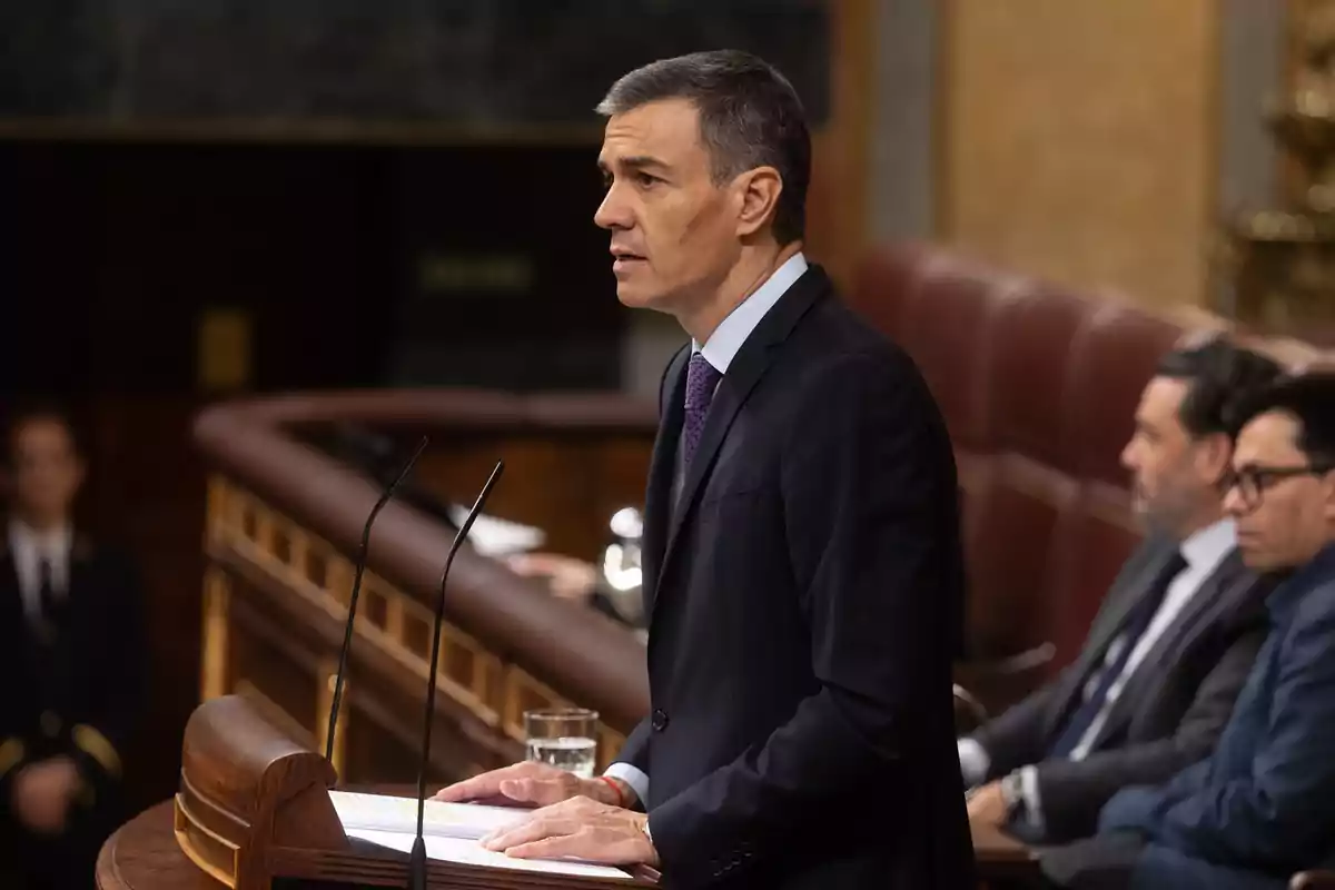 Hombre de traje hablando en un podio en un recinto parlamentario con otras personas sentadas al fondo