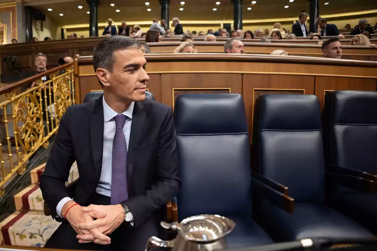 Hombre con traje sentado en un escaño de un parlamento mirando hacia la izquierda mientras otras personas están sentadas al fondo Hombre con traje sentado en un escaño de un parlamento mirando hacia la izquierda mientras otras personas están sentadas al fondo