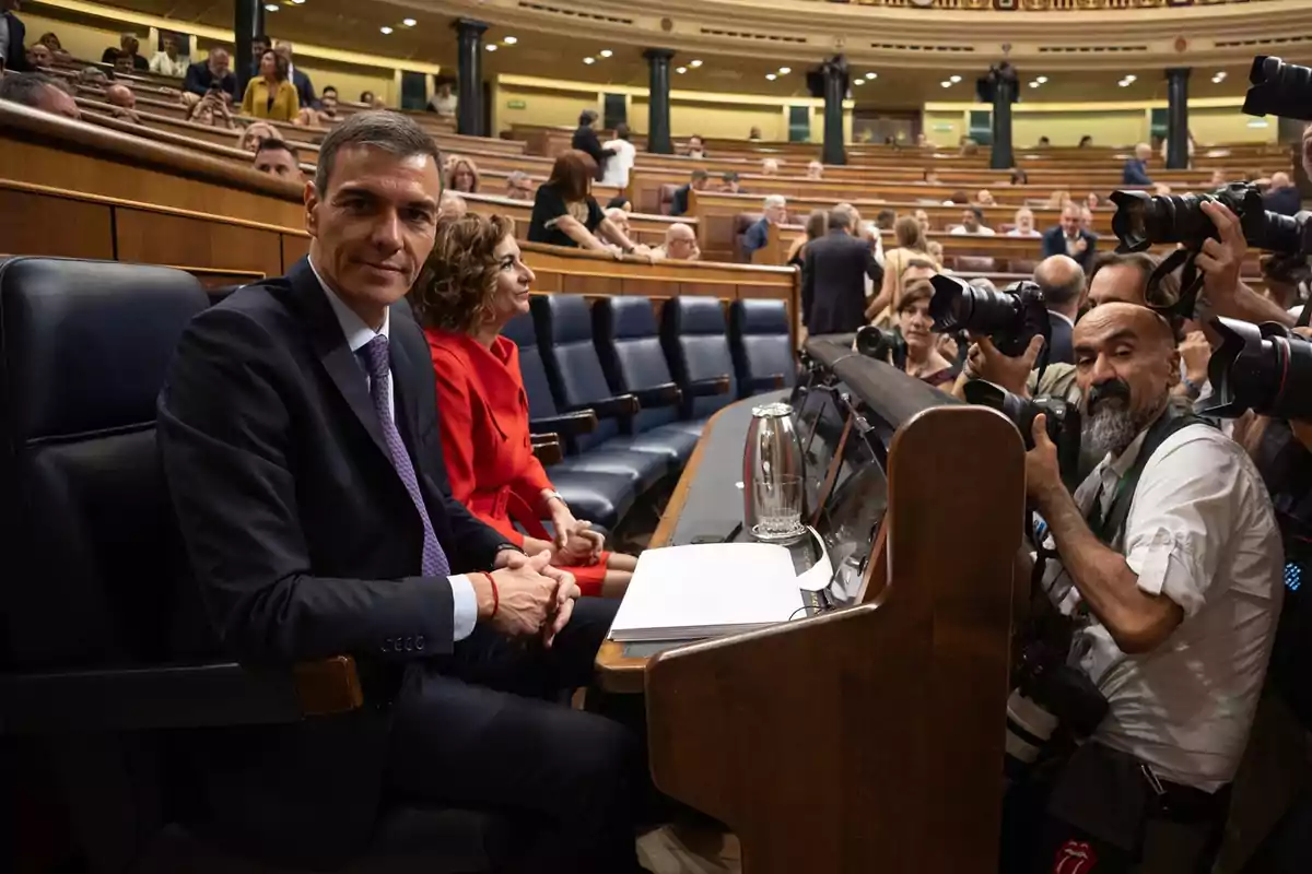 Un hombre con traje y una mujer con vestido rojo están sentados en un hemiciclo mientras varios fotógrafos les toman fotos y otras personas conversan al fondo Un hombre con traje y una mujer con vestido rojo están sentados en un hemiciclo mientras varios fotógrafos les toman fotos y otras personas conversan al fondo