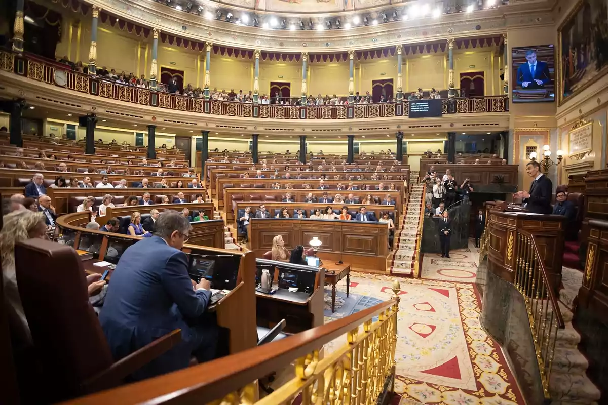 Vista general del interior de un parlamento con numerosos diputados sentados en sus escaños mientras una persona interviene desde la tribuna Vista general del interior de un parlamento con numerosos diputados sentados en sus escaños mientras una persona interviene desde la tribuna