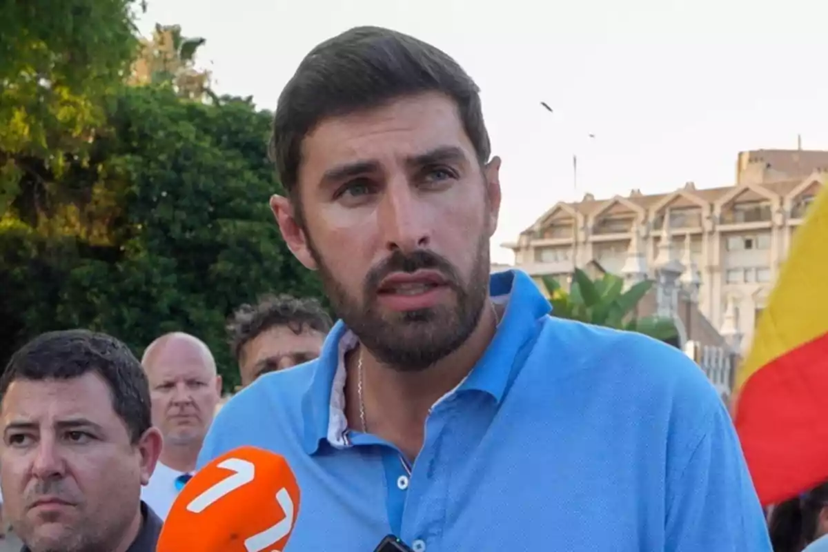 Un hombre con barba y camisa azul habla frente a un micrófono naranja mientras otras personas y una bandera se ven al fondo en un entorno urbano al aire libre Un hombre con barba y camisa azul habla frente a un micrófono naranja mientras otras personas y una bandera se ven al fondo en un entorno urbano al aire libre