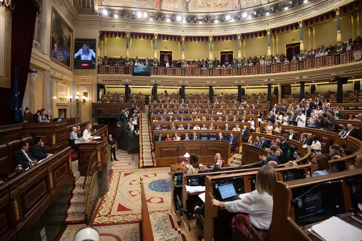 Vista panorámica del interior de un parlamento con numerosos diputados sentados y algunas personas de pie aplaudiendo mientras una oradora habla desde el estrado principal