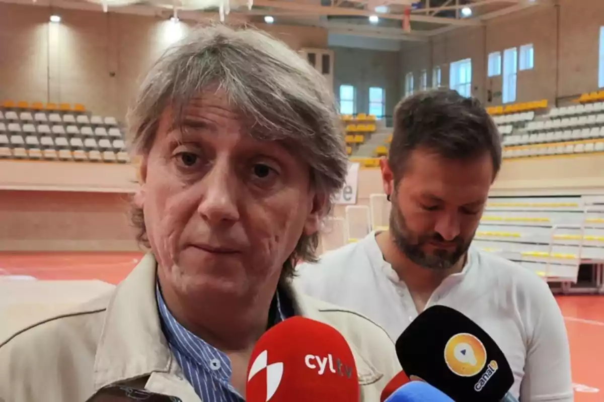 Un hombre de cabello canoso responde preguntas de la prensa en un polideportivo mientras otro hombre está detrás de él mirando hacia abajo Un hombre de cabello canoso responde preguntas de la prensa en un polideportivo mientras otro hombre está detrás de él mirando hacia abajo