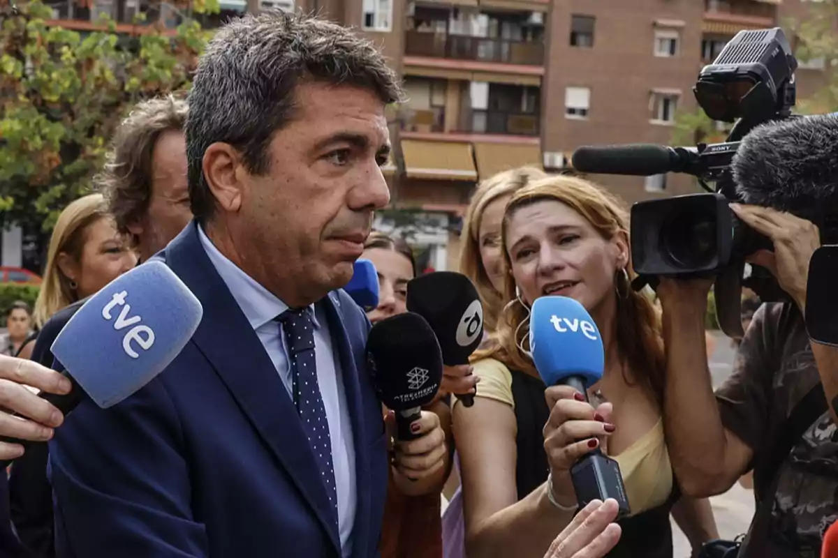 Un hombre con traje azul es rodeado por periodistas y cámaras de televisión mientras responde preguntas en la calle Un hombre con traje azul es rodeado por periodistas y cámaras de televisión mientras responde preguntas en la calle