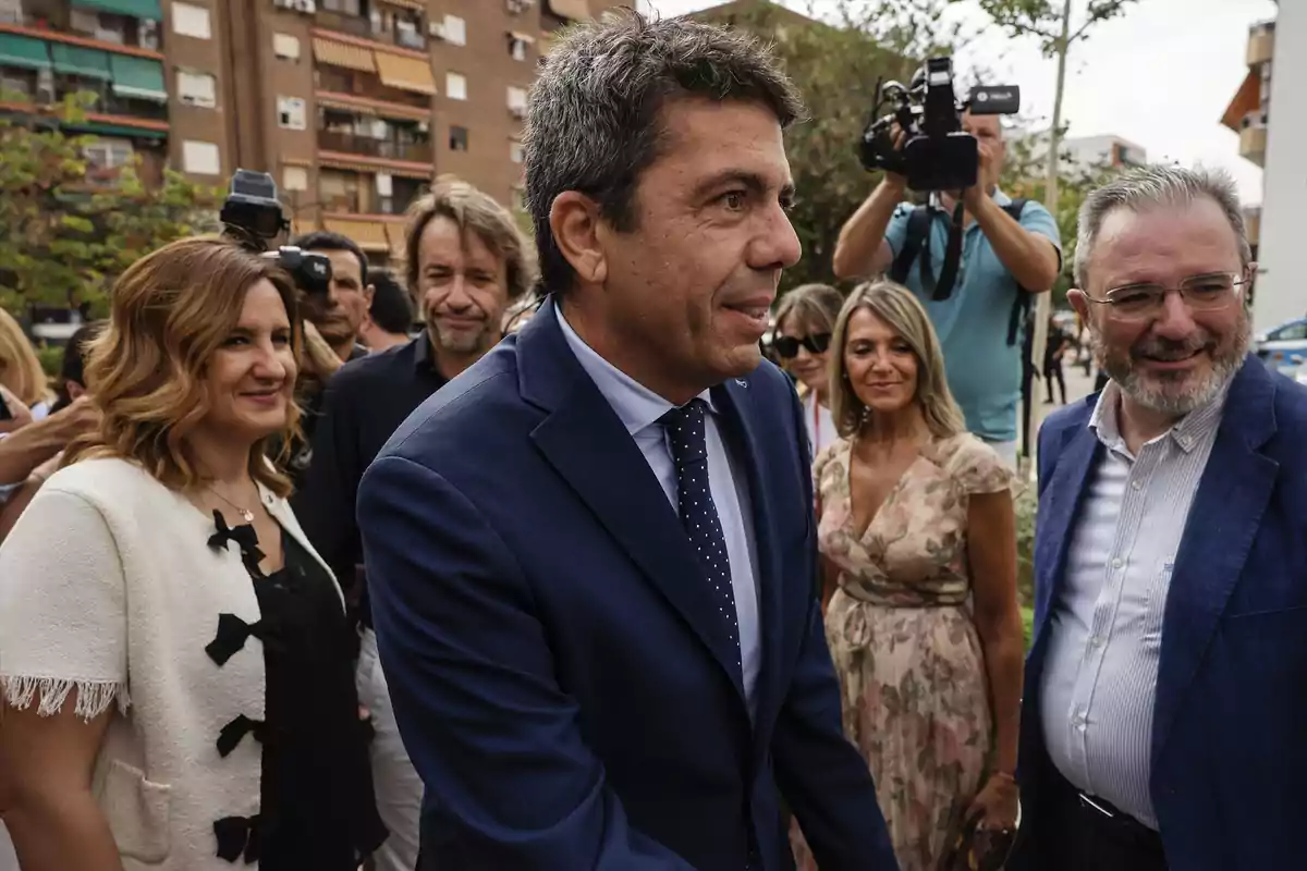 Un hombre con traje azul y corbata saluda mientras está rodeado de varias personas sonrientes y cámaras en una calle urbana