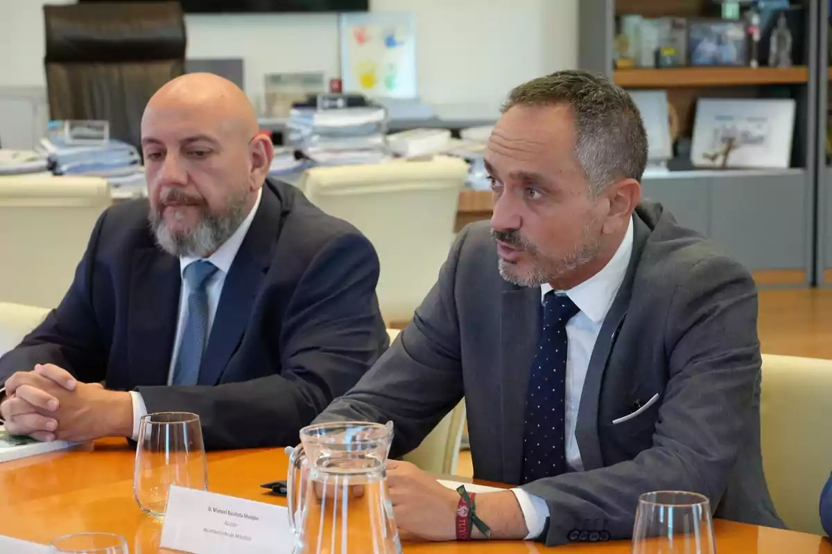 Dos hombres con traje sentados en una mesa de reuniones conversando en una oficina moderna con documentos y vasos de agua sobre la mesa Dos hombres con traje sentados en una mesa de reuniones conversando en una oficina moderna con documentos y vasos de agua sobre la mesa