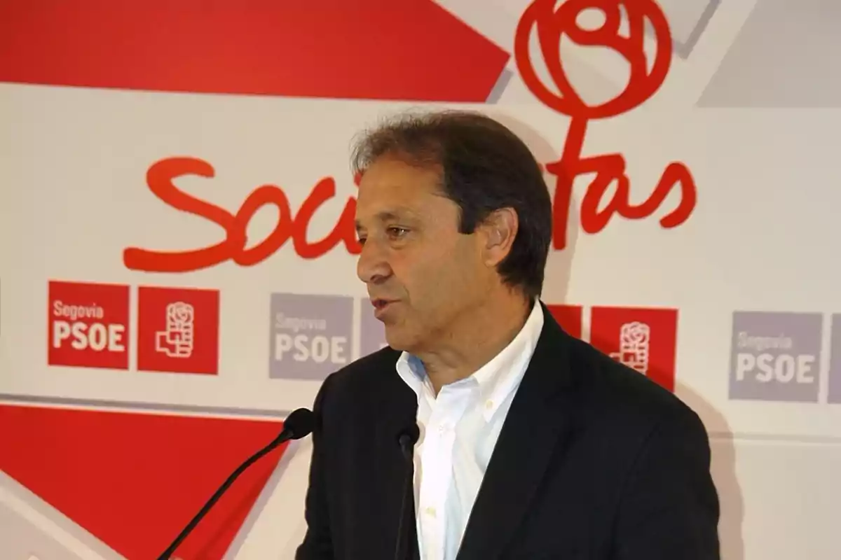 Hombre de mediana edad hablando en un podio con fondo de logotipos del PSOE y la palabra socialistas