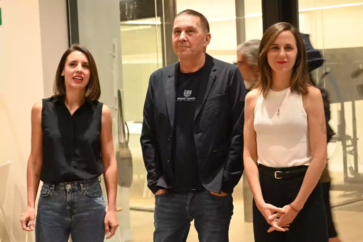 Tres personas posan de pie en un espacio interior iluminado, dos mujeres a los lados y un hombre en el centro, todos mirando hacia el frente y sonriendo levemente. Tres personas posan de pie en un espacio interior iluminado, dos mujeres a los lados y un hombre en el centro, todos mirando hacia el frente y sonriendo levemente.