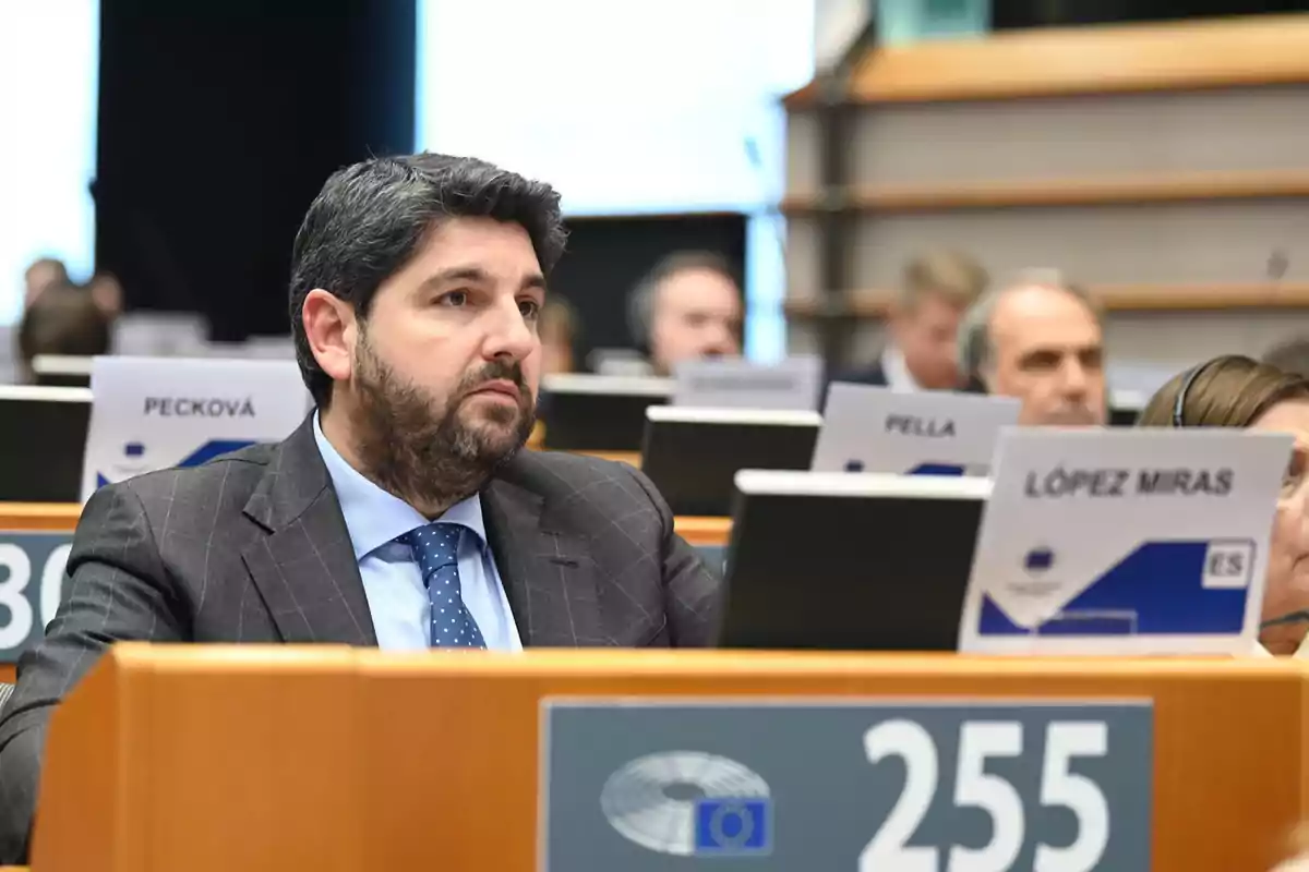 Hombre de traje sentado en una sala de conferencias con otros asistentes y carteles con nombres en los escritorios Hombre de traje sentado en una sala de conferencias con otros asistentes y carteles con nombres en los escritorios