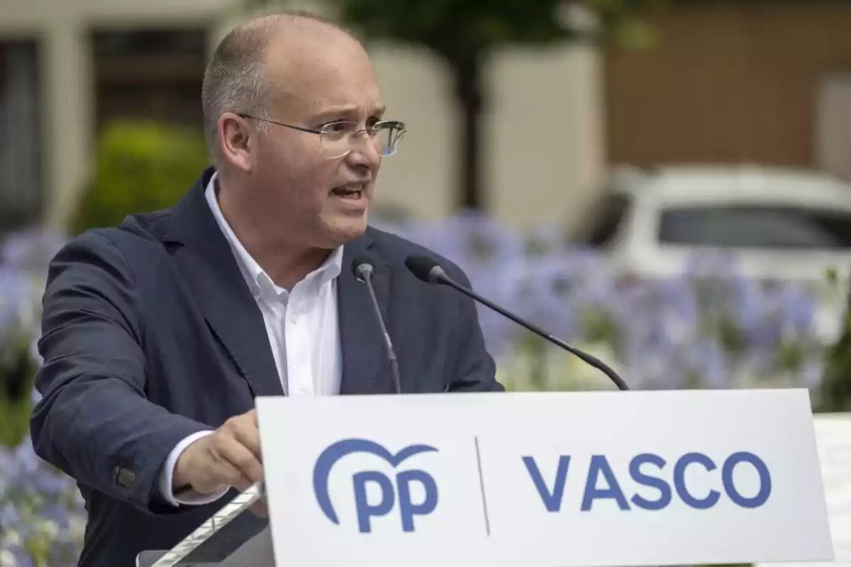 Hombre con gafas y chaqueta azul hablando en un mitin al aire libre frente a un atril con el logo de PP Vasco