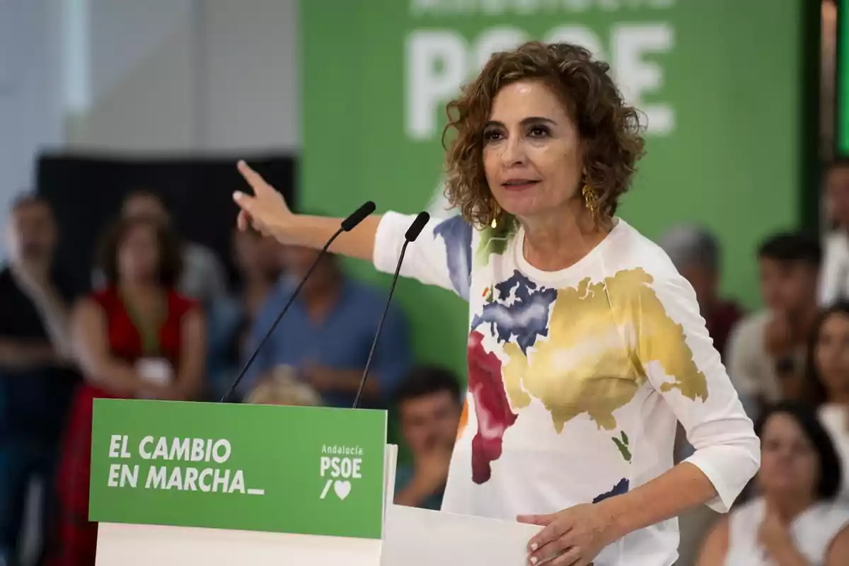 Mujer de cabello rizado hablando en un mitin político del PSOE en Andalucía señalando hacia un lado con un fondo verde y personas al fondo