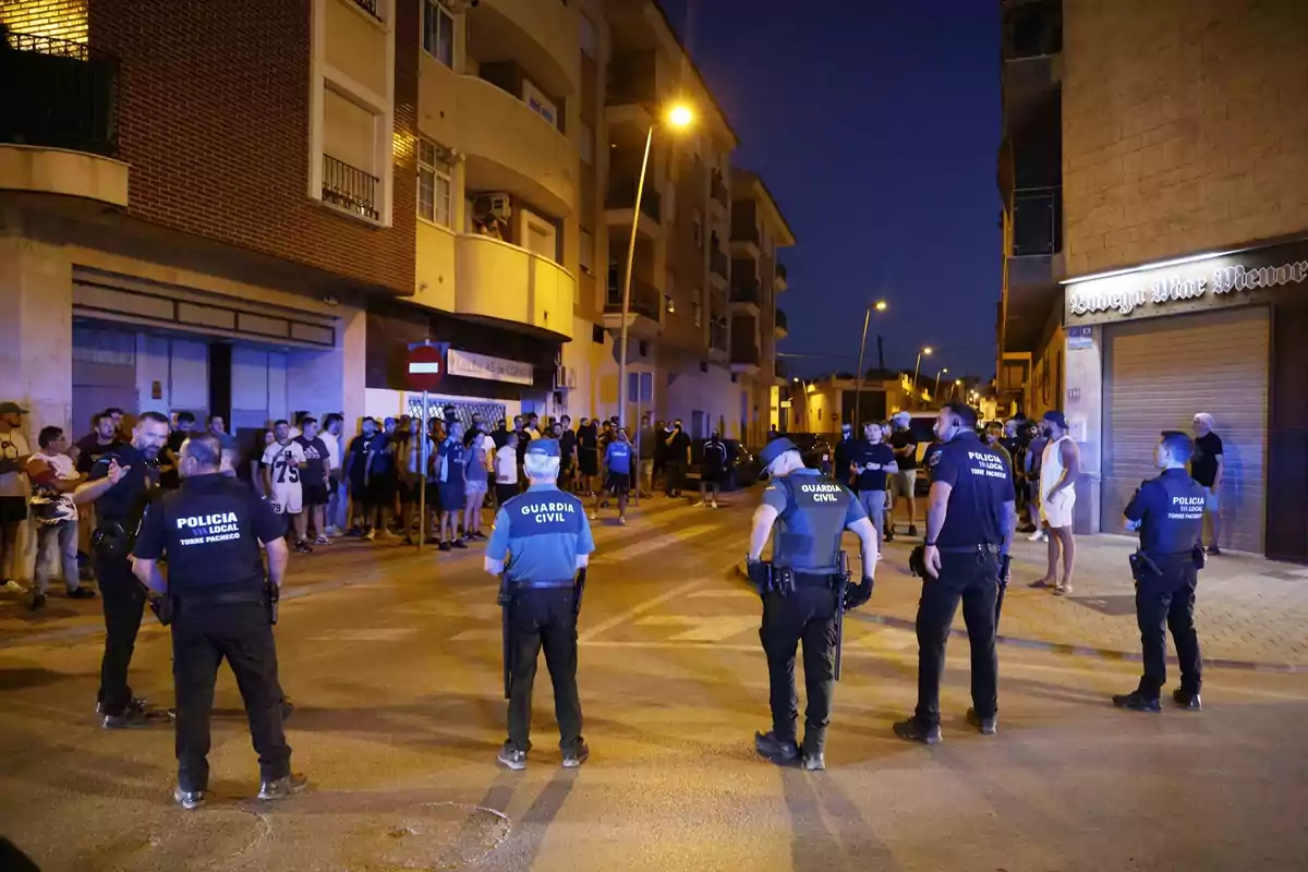 Varios agentes de policía y guardia civil forman un cordón de seguridad frente a un grupo de personas en una calle de noche iluminada por farolas Varios agentes de policía y guardia civil forman un cordón de seguridad frente a un grupo de personas en una calle de noche iluminada por farolas