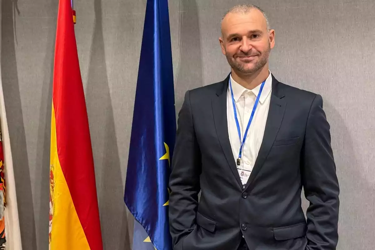 Hombre con traje oscuro y gafete posando junto a las banderas de España y la Unión Europea Hombre con traje oscuro y gafete posando junto a las banderas de España y la Unión Europea