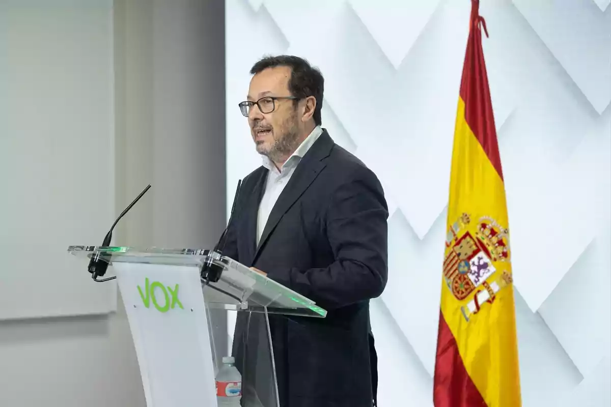 Hombre con gafas hablando en un atril de VOX junto a una bandera de España