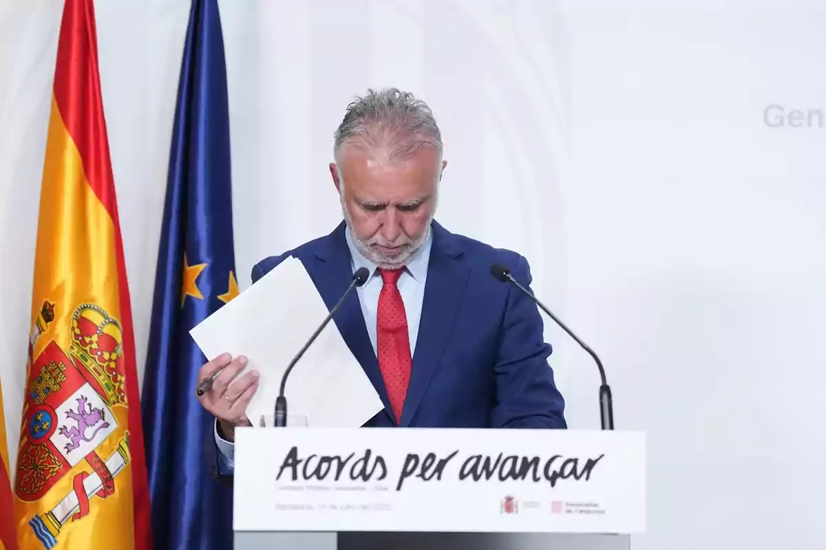 Hombre de cabello canoso y barba con traje azul y corbata roja sostiene unos papeles mientras está de pie frente a un atril con el texto Acords per avançar y dos micrófonos, junto a las banderas de España y la Unión Europea. Hombre de cabello canoso y barba con traje azul y corbata roja sostiene unos papeles mientras está de pie frente a un atril con el texto Acords per avançar y dos micrófonos, junto a las banderas de España y la Unión Europea.