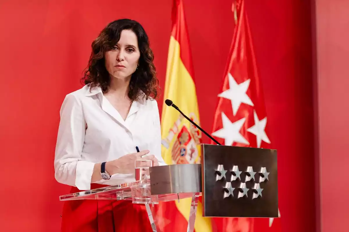 Mujer de cabello rizado y camisa blanca hablando en un atril con fondo rojo y banderas de España y la Comunidad de Madrid