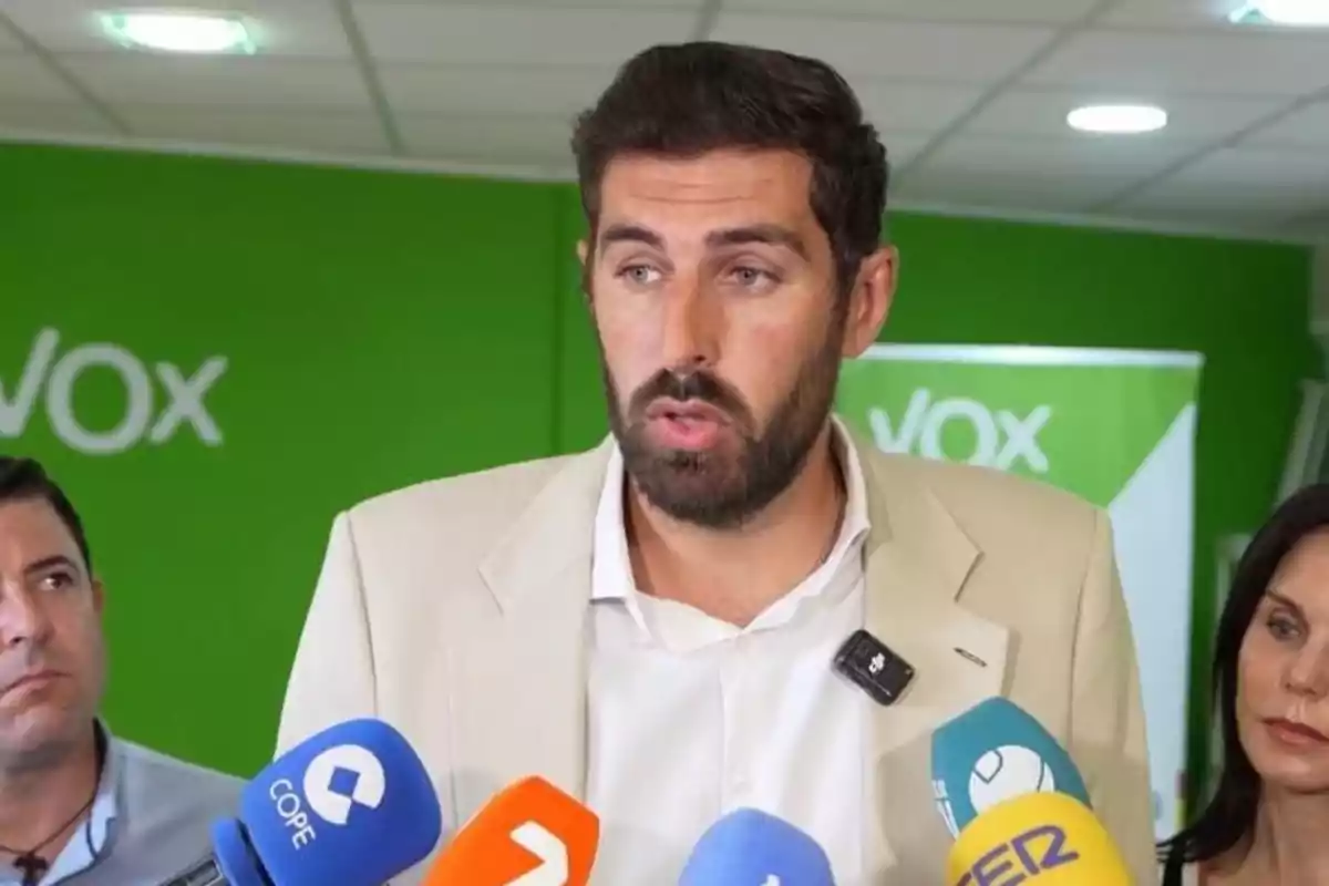Un hombre con barba y chaqueta clara habla frente a varios micrófonos de diferentes medios de comunicación con un fondo verde donde se lee parte del logo de Vox