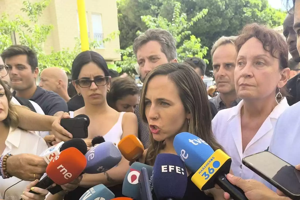 Una mujer rodeada de periodistas responde preguntas frente a varios micrófonos de diferentes medios de comunicación mientras otras personas la observan atentamente