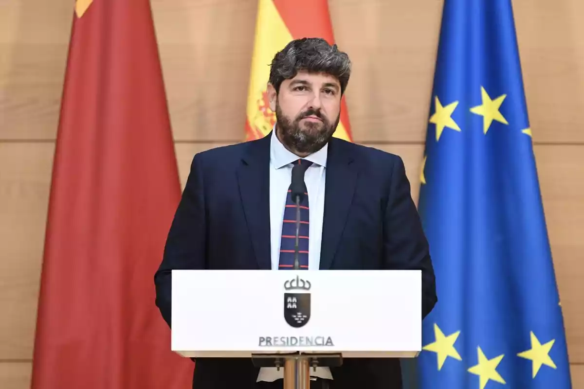 Hombre con barba y traje oscuro hablando en un atril de presidencia con banderas de España, la Unión Europea y una bandera roja de fondo