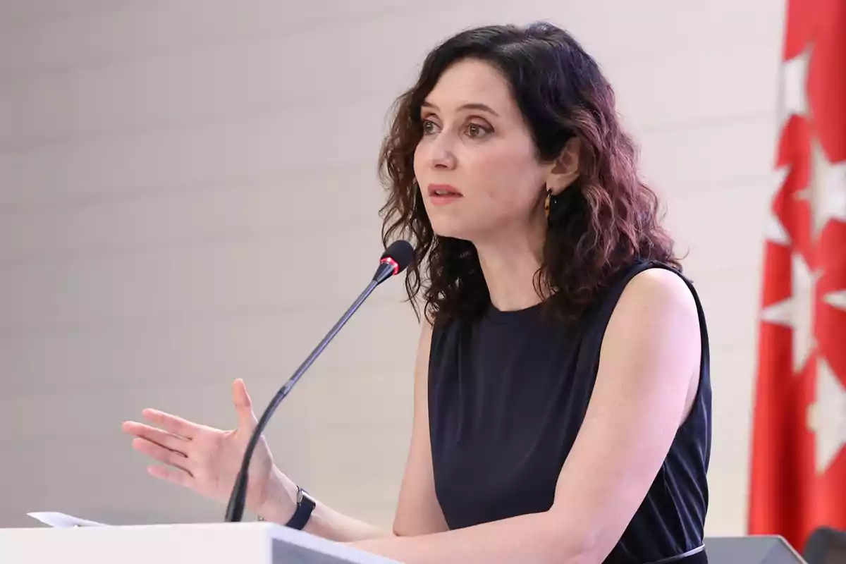 Mujer de cabello rizado y oscuro hablando en un micrófono durante una conferencia con una bandera roja al fondo