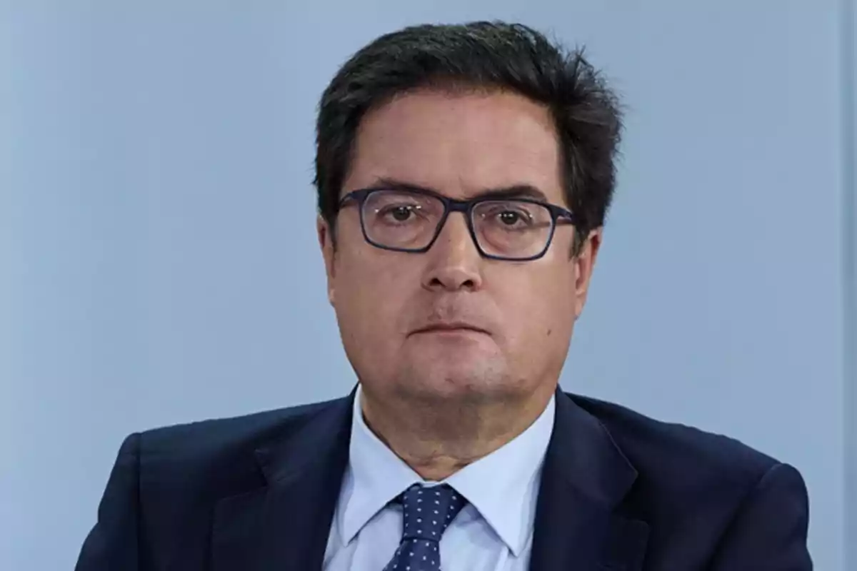 Hombre de mediana edad con gafas y traje oscuro mirando al frente con expresión seria sobre un fondo azul claro Hombre de mediana edad con gafas y traje oscuro mirando al frente con expresión seria sobre un fondo azul claro