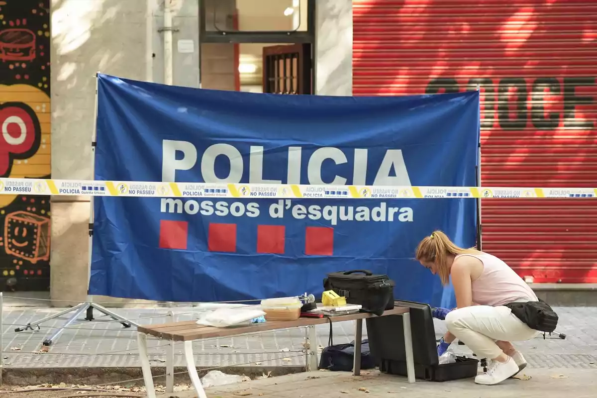 Una agente trabaja en la escena de un crimen acordonada por la policía en una calle urbana, con un cartel azul de los Mossos d'Esquadra de fondo y material de investigación sobre un banco. Una agente trabaja en la escena de un crimen acordonada por la policía en una calle urbana, con un cartel azul de los Mossos d'Esquadra de fondo y material de investigación sobre un banco.
