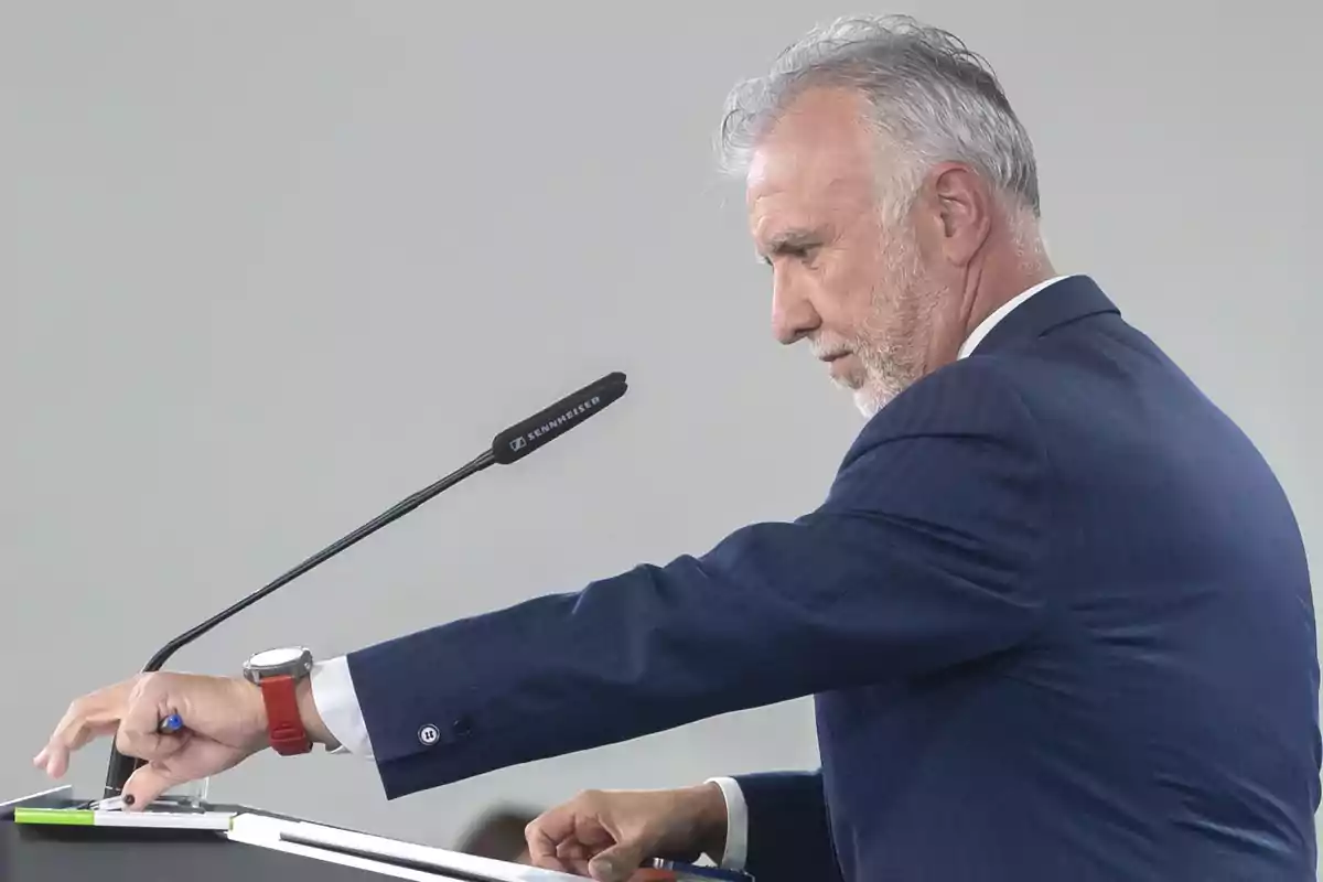 Hombre de cabello canoso y barba vestido con traje azul hablando en un podio con micrófono y tomando notas Hombre de cabello canoso y barba vestido con traje azul hablando en un podio con micrófono y tomando notas