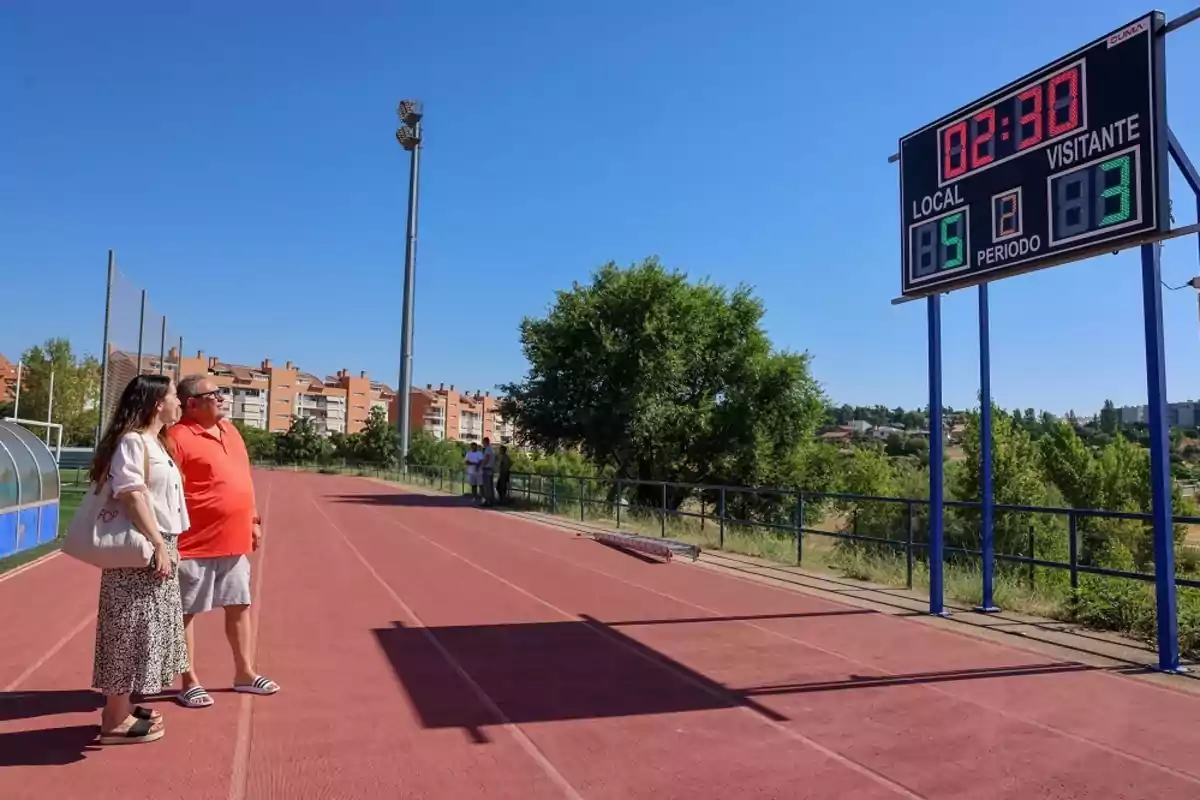 Dos personas observan un marcador electrónico en una pista de atletismo al aire libre en un día soleado