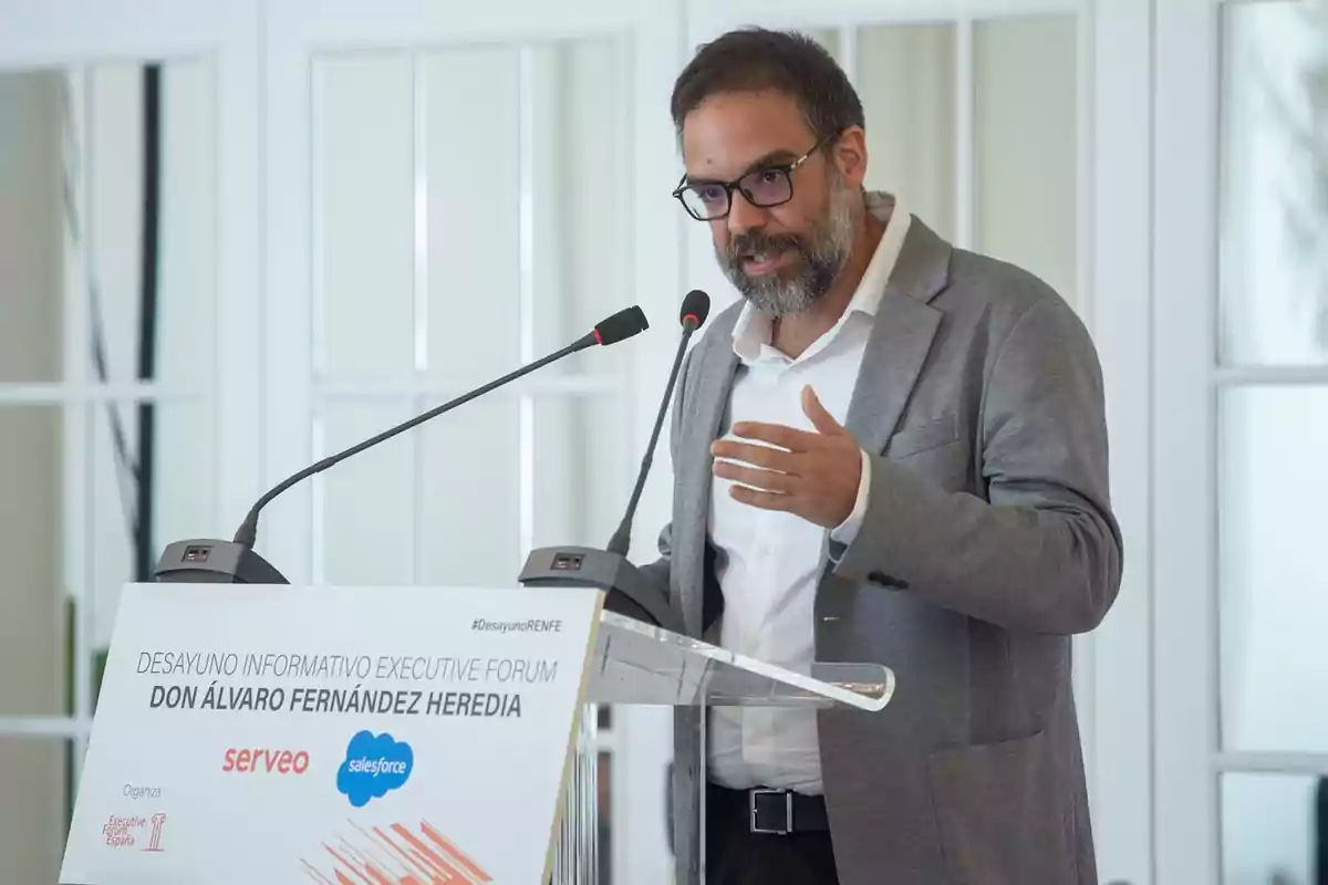 Hombre con gafas y barba hablando en un podio durante un evento ejecutivo, vistiendo saco gris y camisa blanca Hombre con gafas y barba hablando en un podio durante un evento ejecutivo, vistiendo saco gris y camisa blanca
