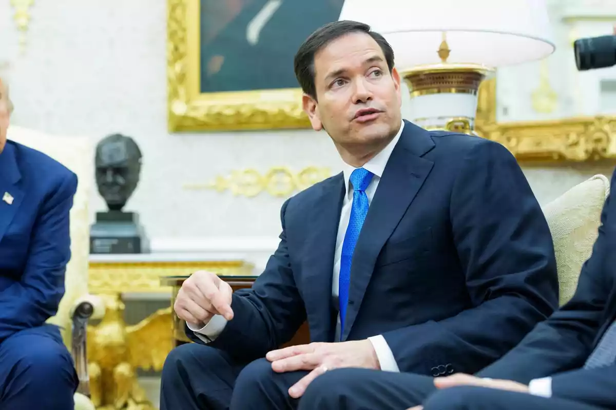 Hombre de traje azul y corbata azul sentado en una sala elegante mientras conversa con otras personas Hombre de traje azul y corbata azul sentado en una sala elegante mientras conversa con otras personas