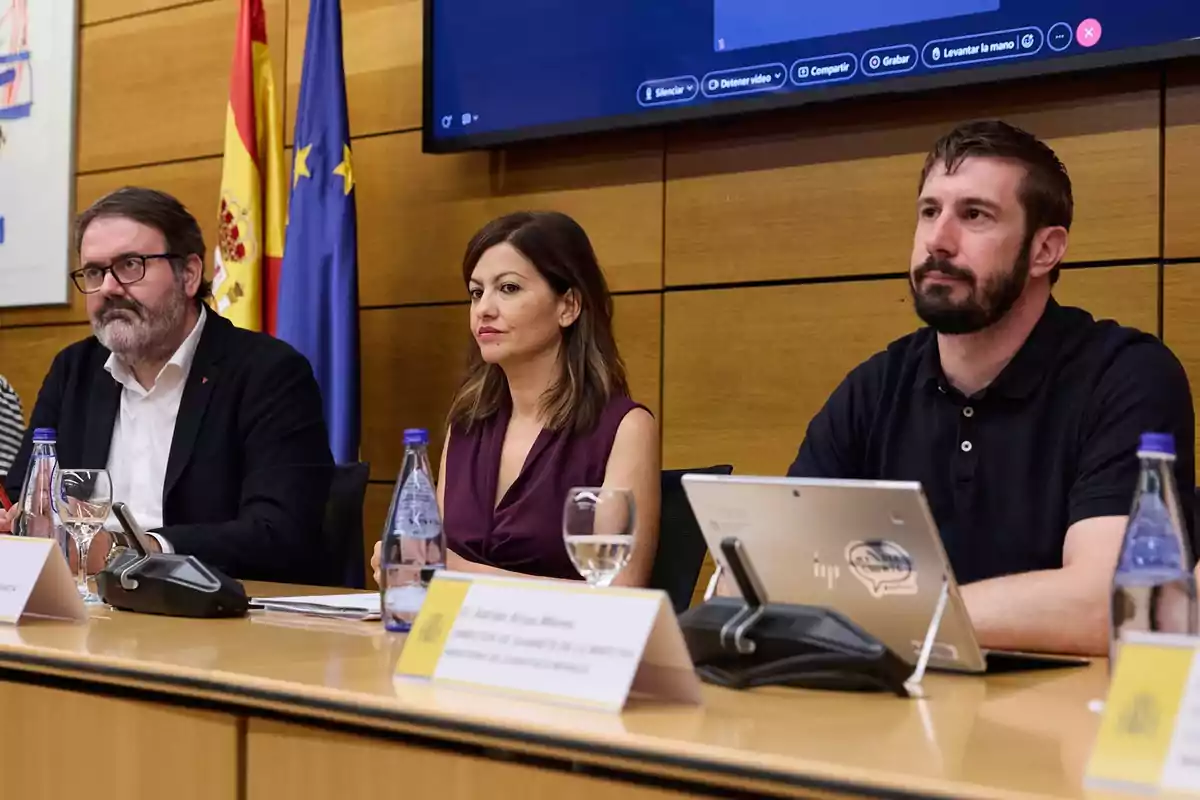 Tres personas sentadas en una mesa de conferencia con botellas de agua, copas y dispositivos electrónicos, al fondo hay banderas de España y la Unión Europea y una pantalla grande en la pared. Tres personas sentadas en una mesa de conferencia con botellas de agua, copas y dispositivos electrónicos, al fondo hay banderas de España y la Unión Europea y una pantalla grande en la pared.