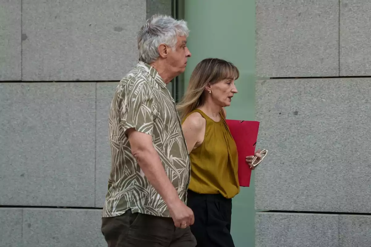 Dos personas caminan juntas por la acera frente a una pared gris, una mujer lleva una carpeta roja y un hombre viste una camisa de estampado vegetal. Dos personas caminan juntas por la acera frente a una pared gris, una mujer lleva una carpeta roja y un hombre viste una camisa de estampado vegetal.