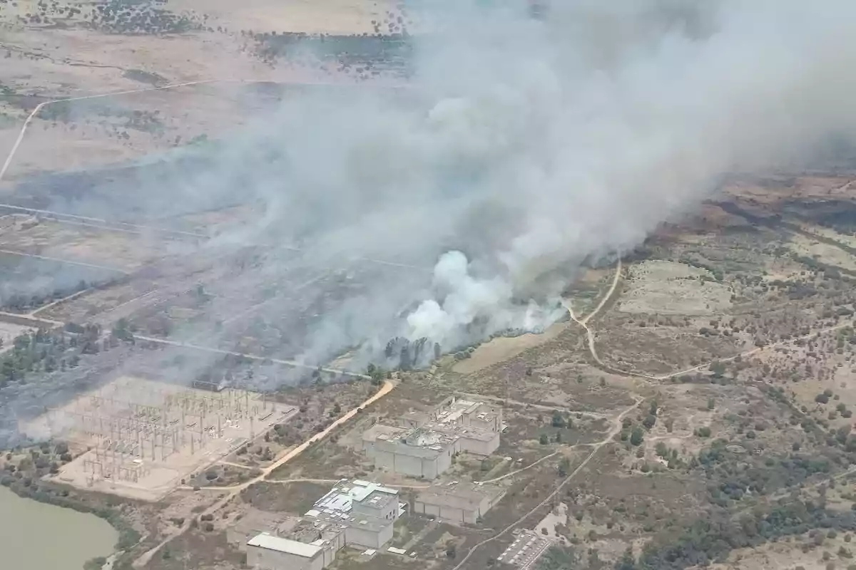 Vista aérea de un incendio forestal que genera una gran columna de humo cerca de una subestación eléctrica y varios edificios en una zona rural