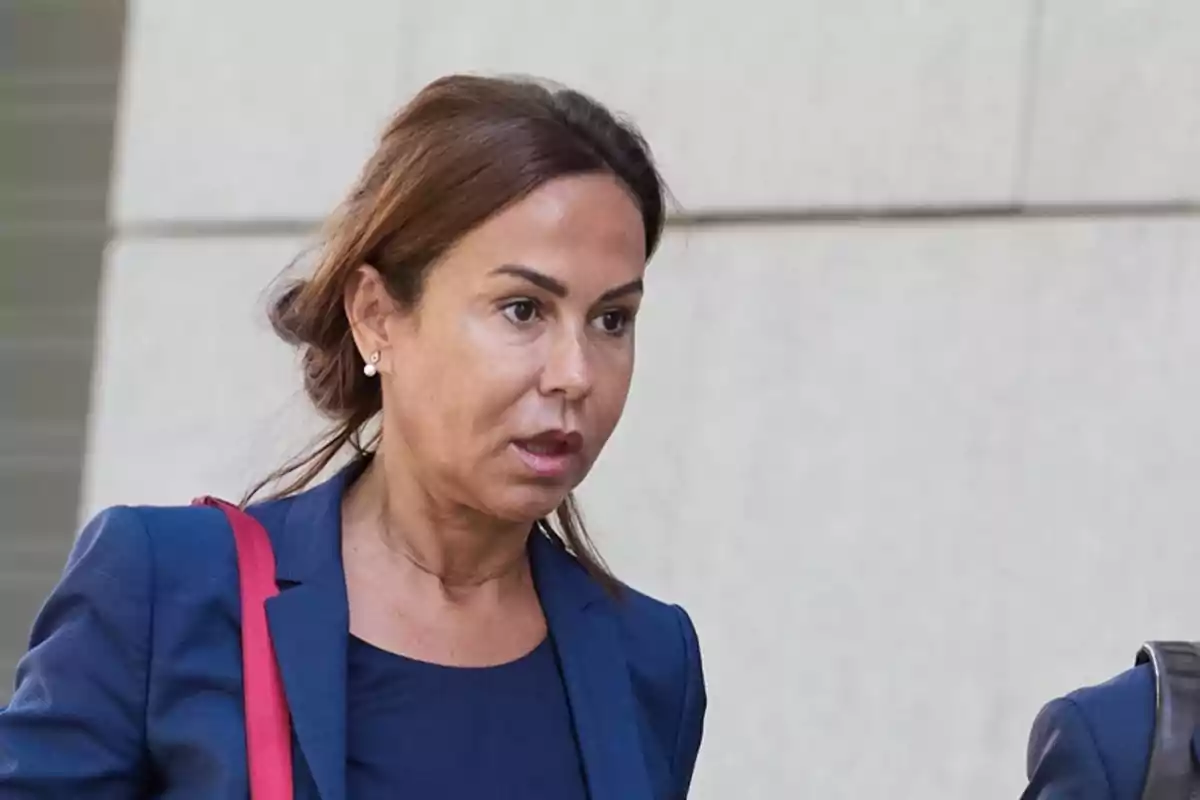 Mujer de cabello castaño recogido vestida con saco azul y blusa azul oscuro lleva una bolsa roja y camina frente a una pared clara