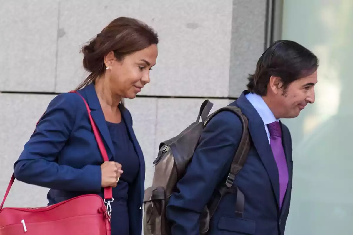 Dos personas vestidas de traje caminan juntas, una mujer lleva un bolso rojo y un hombre lleva una mochila negra.