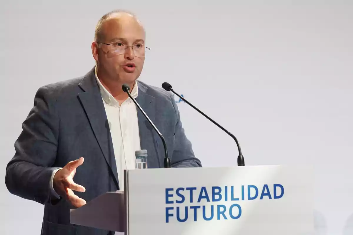 Hombre con gafas y saco azul hablando en un podio con dos micrófonos y un cartel que dice ESTABILIDAD FUTURO Hombre con gafas y saco azul hablando en un podio con dos micrófonos y un cartel que dice ESTABILIDAD FUTURO