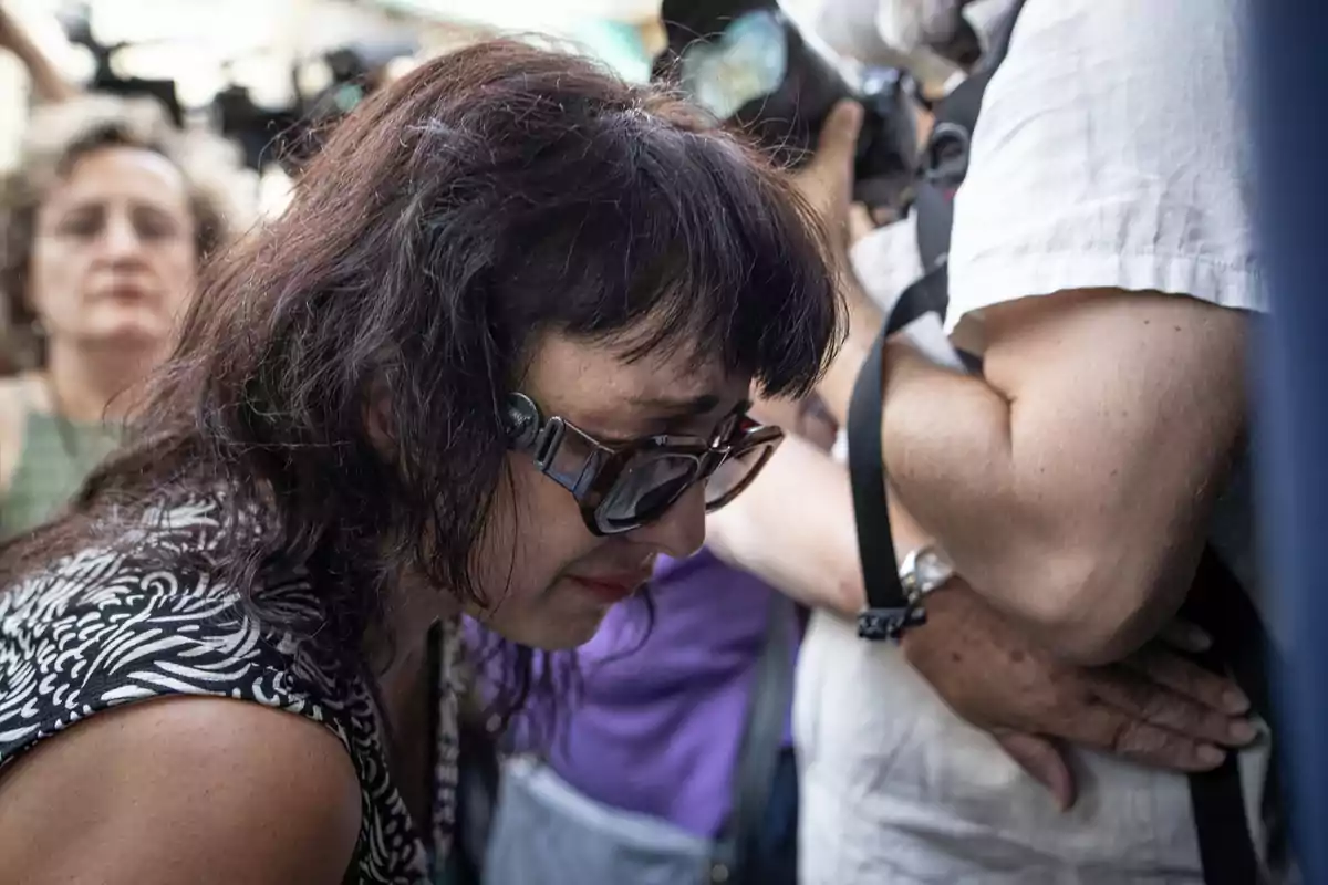 Mujer con gafas oscuras y expresión de tristeza inclinada hacia adelante entre una multitud