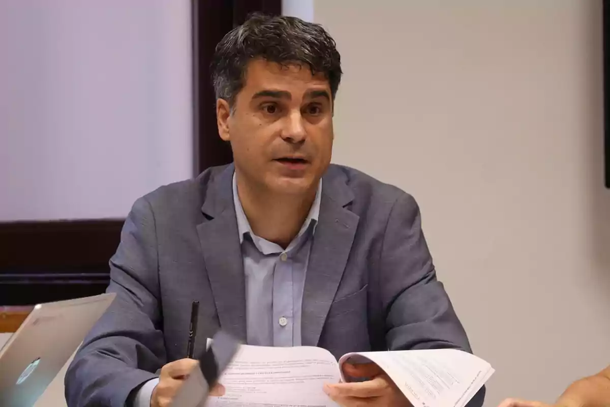 Hombre de cabello oscuro con saco azul claro sentado en una mesa sosteniendo documentos y un bolígrafo mientras parece estar hablando
