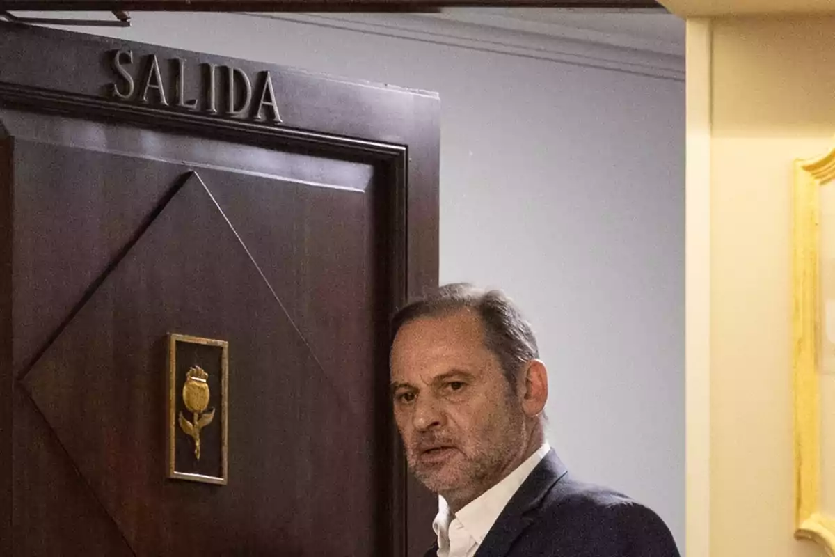 Hombre de cabello canoso y barba frente a una puerta de madera con un letrero que dice salida Hombre de cabello canoso y barba frente a una puerta de madera con un letrero que dice salida