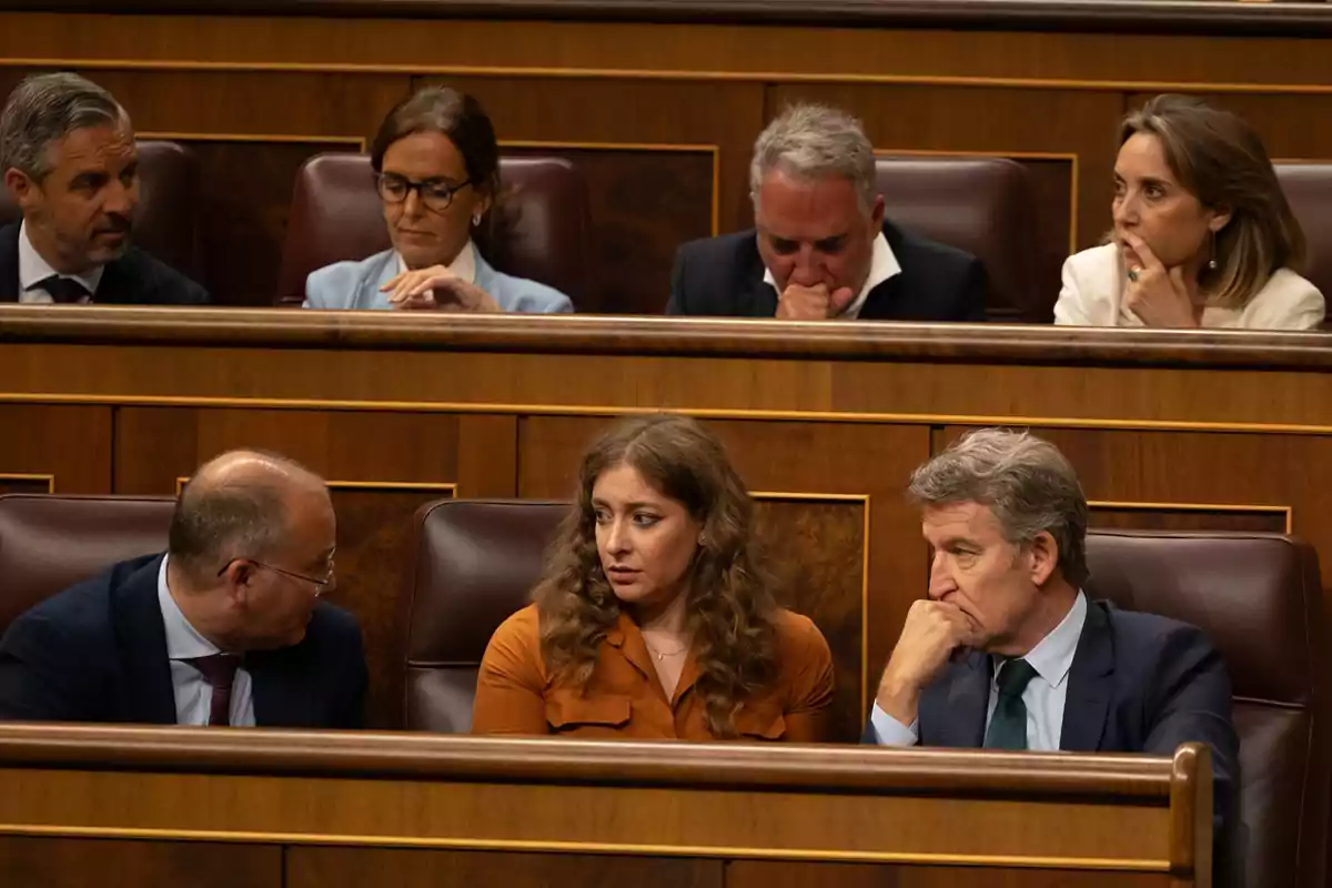 Varias personas sentadas en escaños de madera en lo que parece ser un parlamento, algunas conversando y otras pensativas Varias personas sentadas en escaños de madera en lo que parece ser un parlamento, algunas conversando y otras pensativas