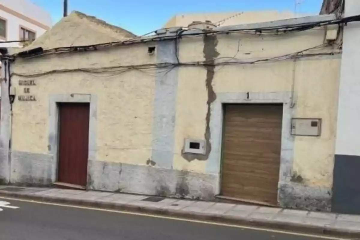 Fachada de una casa antigua con paredes amarillas y detalles grises, dos puertas de madera y varias grietas visibles, situada junto a una acera y una calle asfaltada. Fachada de una casa antigua con paredes amarillas y detalles grises, dos puertas de madera y varias grietas visibles, situada junto a una acera y una calle asfaltada.