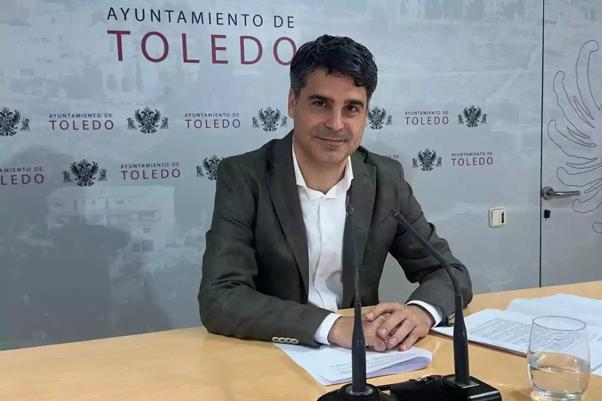 Hombre con cabello oscuro y saco gris sentado en una mesa con micrófonos y papeles frente a un fondo que dice Ayuntamiento de Toledo Hombre con cabello oscuro y saco gris sentado en una mesa con micrófonos y papeles frente a un fondo que dice Ayuntamiento de Toledo