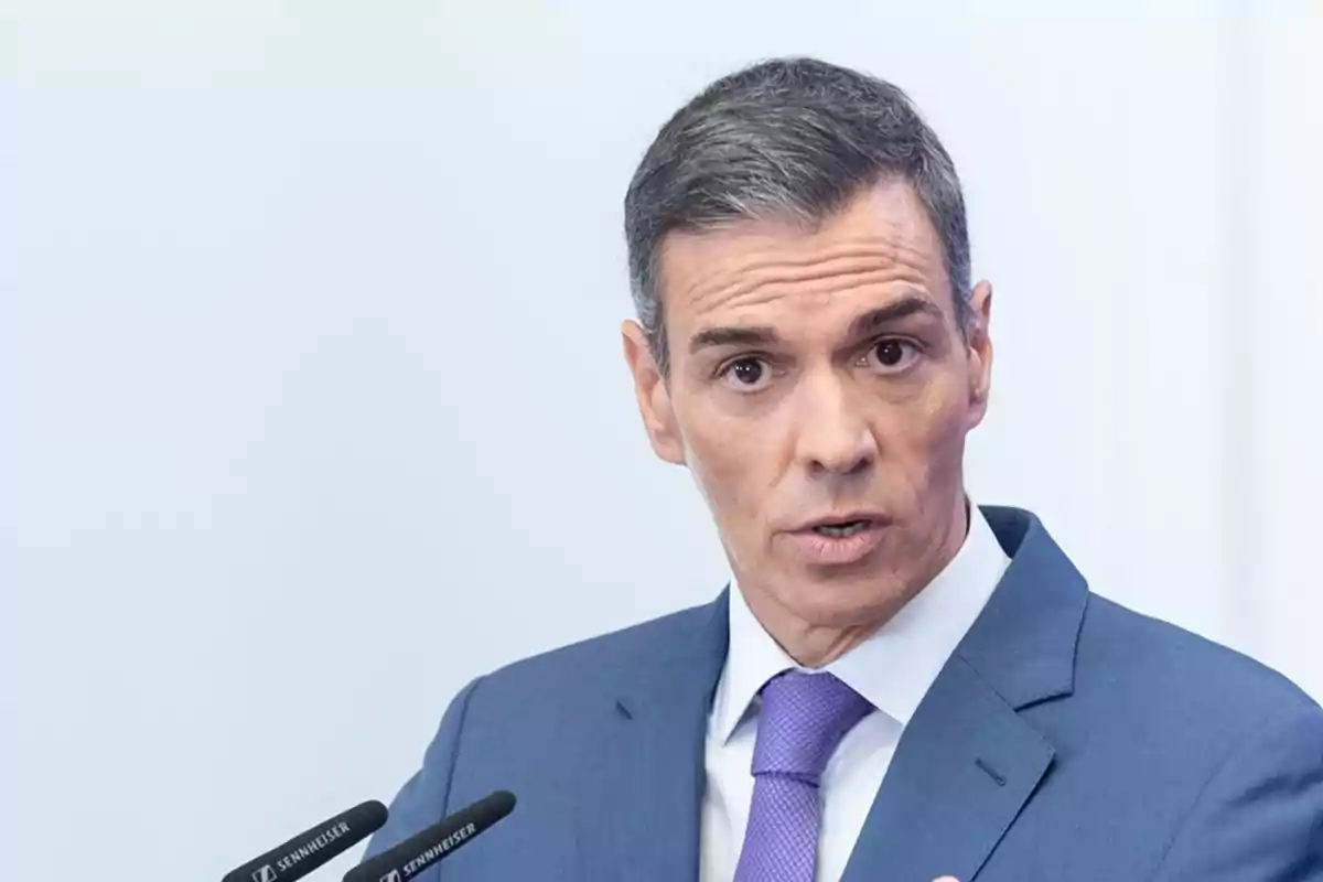 Hombre de cabello corto y canoso con traje azul y corbata morada hablando frente a dos micrófonos Hombre de cabello corto y canoso con traje azul y corbata morada hablando frente a dos micrófonos