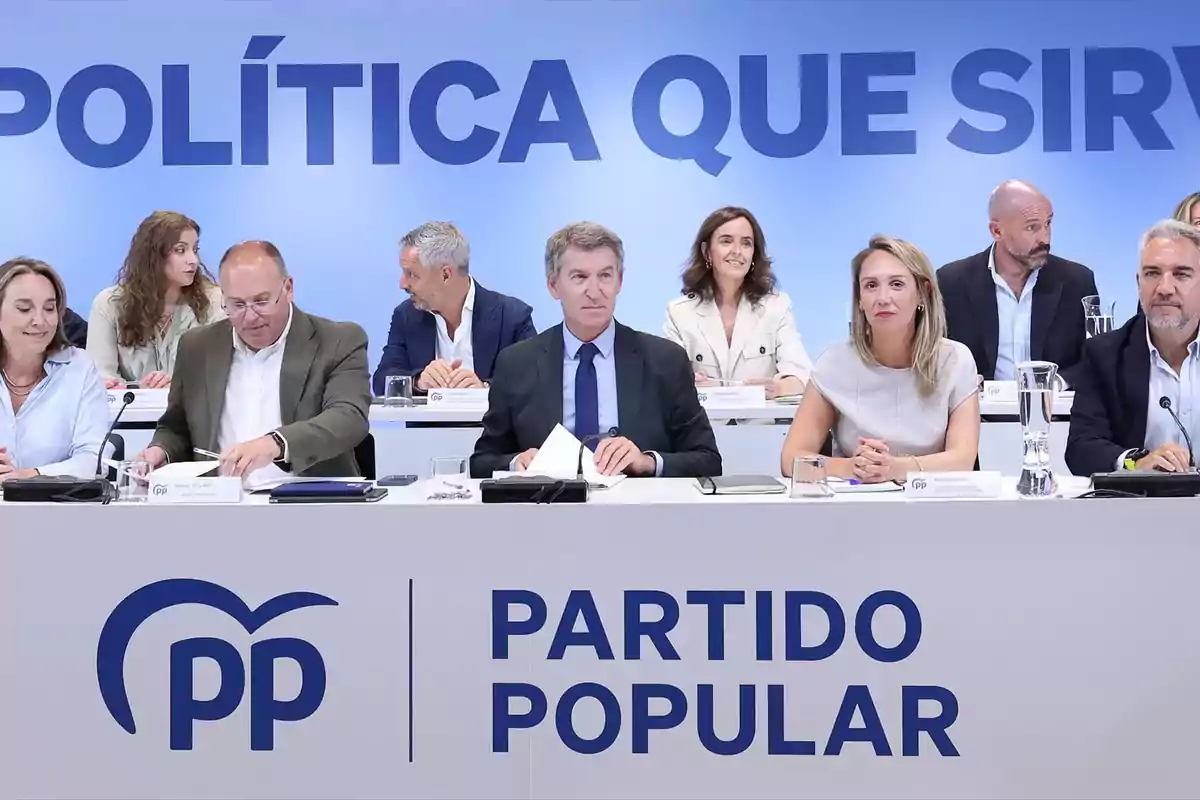 Varias personas sentadas en una mesa durante una reunión del Partido Popular con un cartel de fondo que dice política que sirve