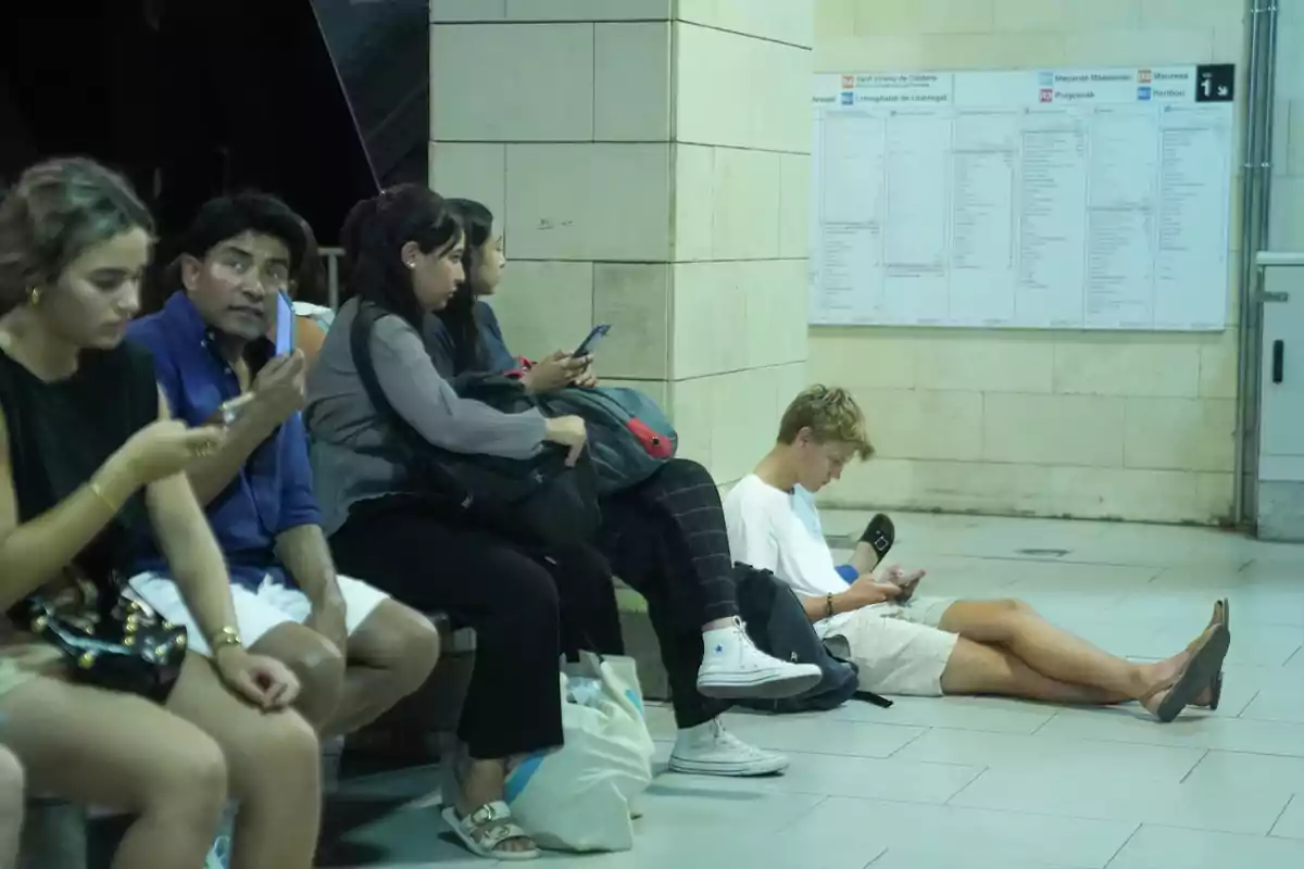 Varias personas esperan sentadas y usando sus teléfonos móviles en una estación de metro Varias personas esperan sentadas y usando sus teléfonos móviles en una estación de metro