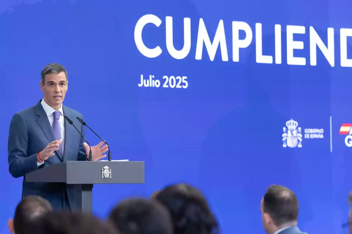 Hombre con traje azul y corbata lila hablando en un atril frente a una pantalla azul que muestra el texto Cumpliendo Julio 2025 y logotipos del Gobierno de España