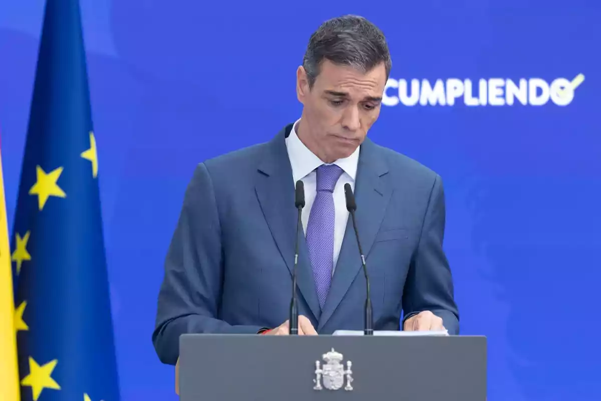 Hombre con traje azul y corbata lila hablando en un podio frente a un fondo azul con banderas y la palabra cumpliendo parcialmente visible
